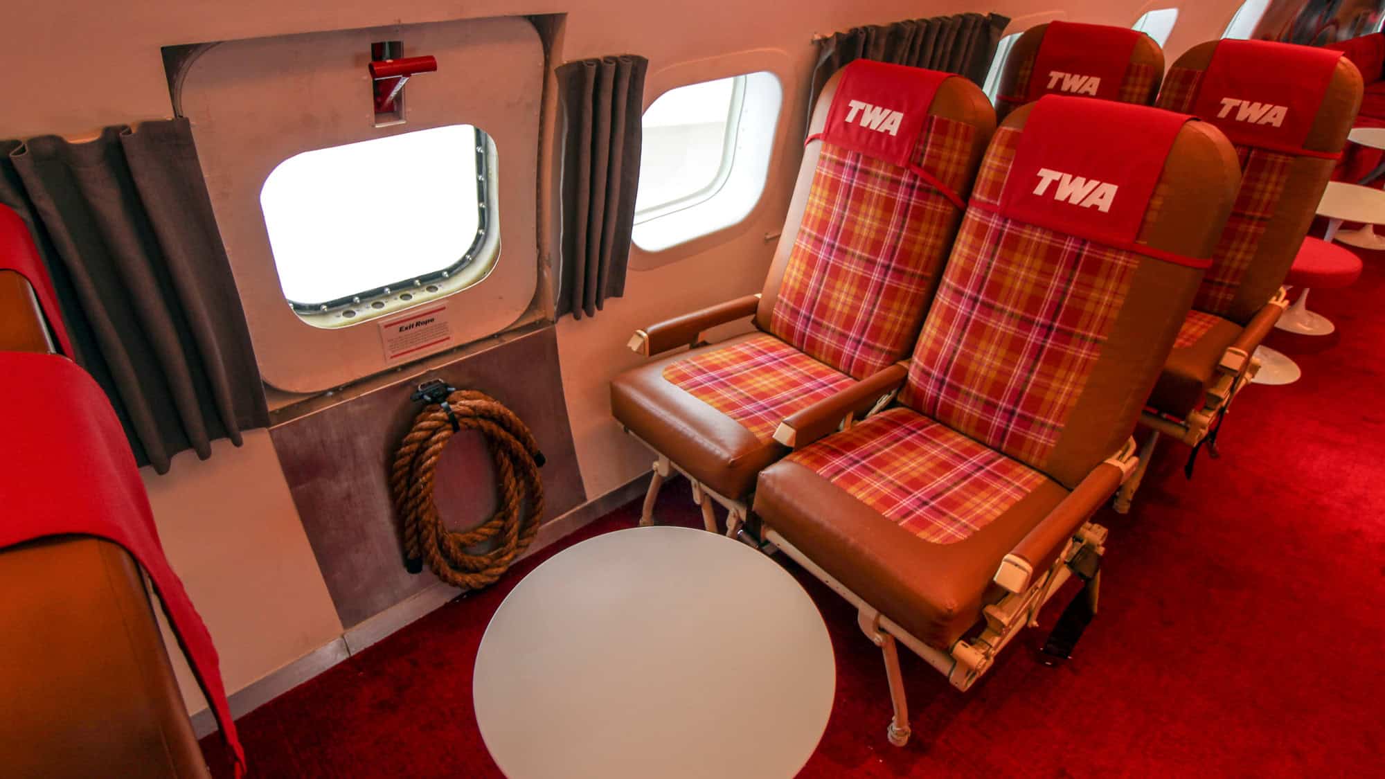 Interior of a retro TWA airplane cabin featuring red and orange plaid seats, red carpeting, and an emergency exit with a coiled rope labeled 'Exit Rope.'