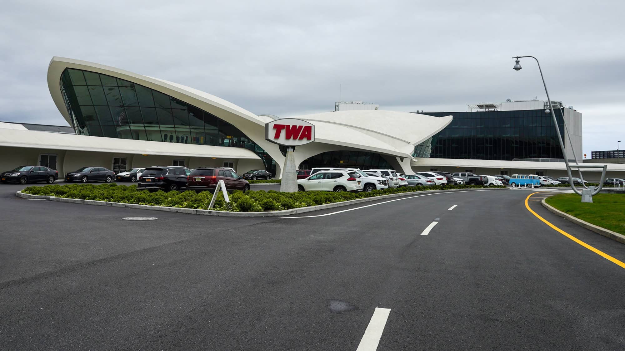 Exterior view of the TWA Flight Center at JFK Airport, showcasing its iconic wing-like roof, a red 'TWA' sign, and a parking area with vintage and modern vehicles.
