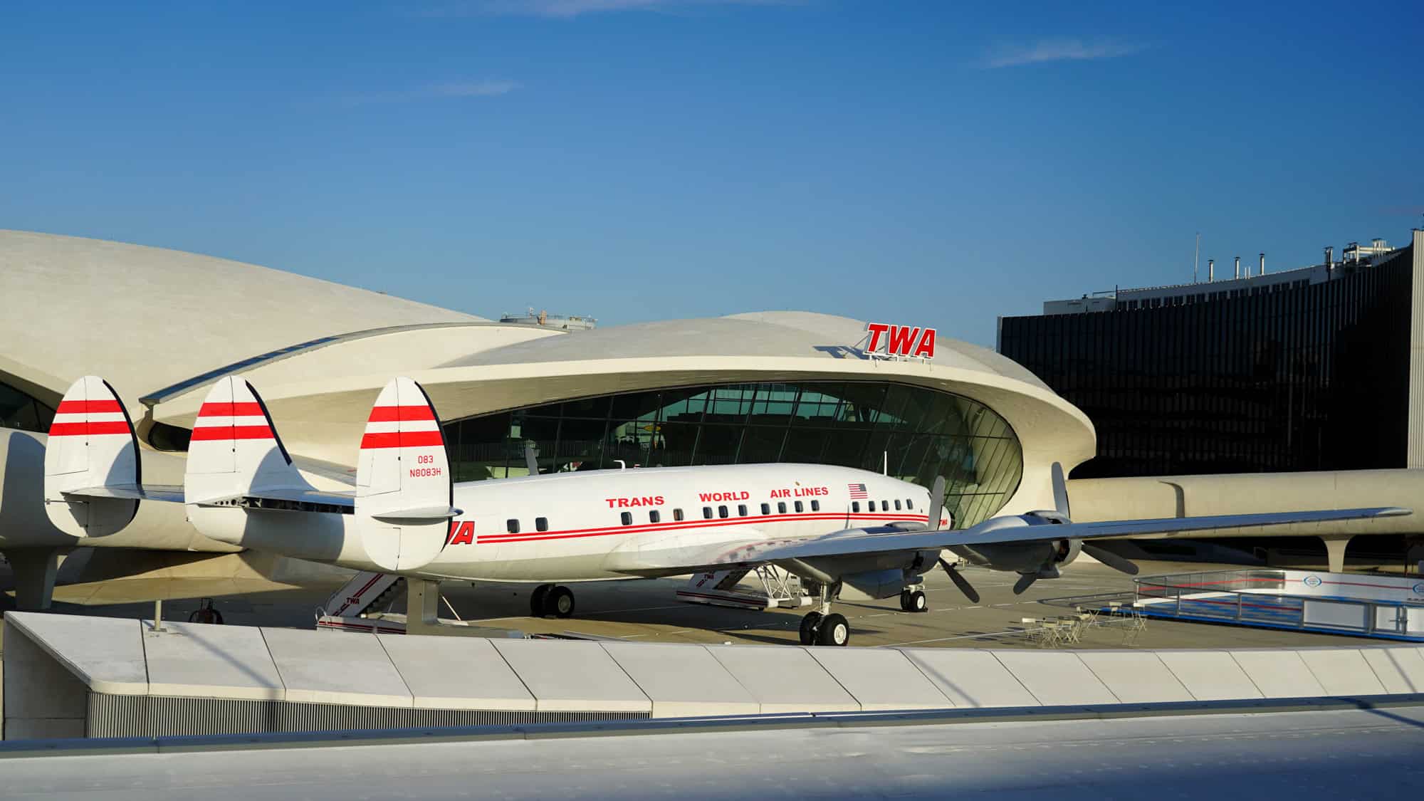 A vintage TWA Lockheed Constellation airplane parked in front of the iconic TWA Hotel terminal, with its distinctive triple tailfins and red branding under a clear blue sky.