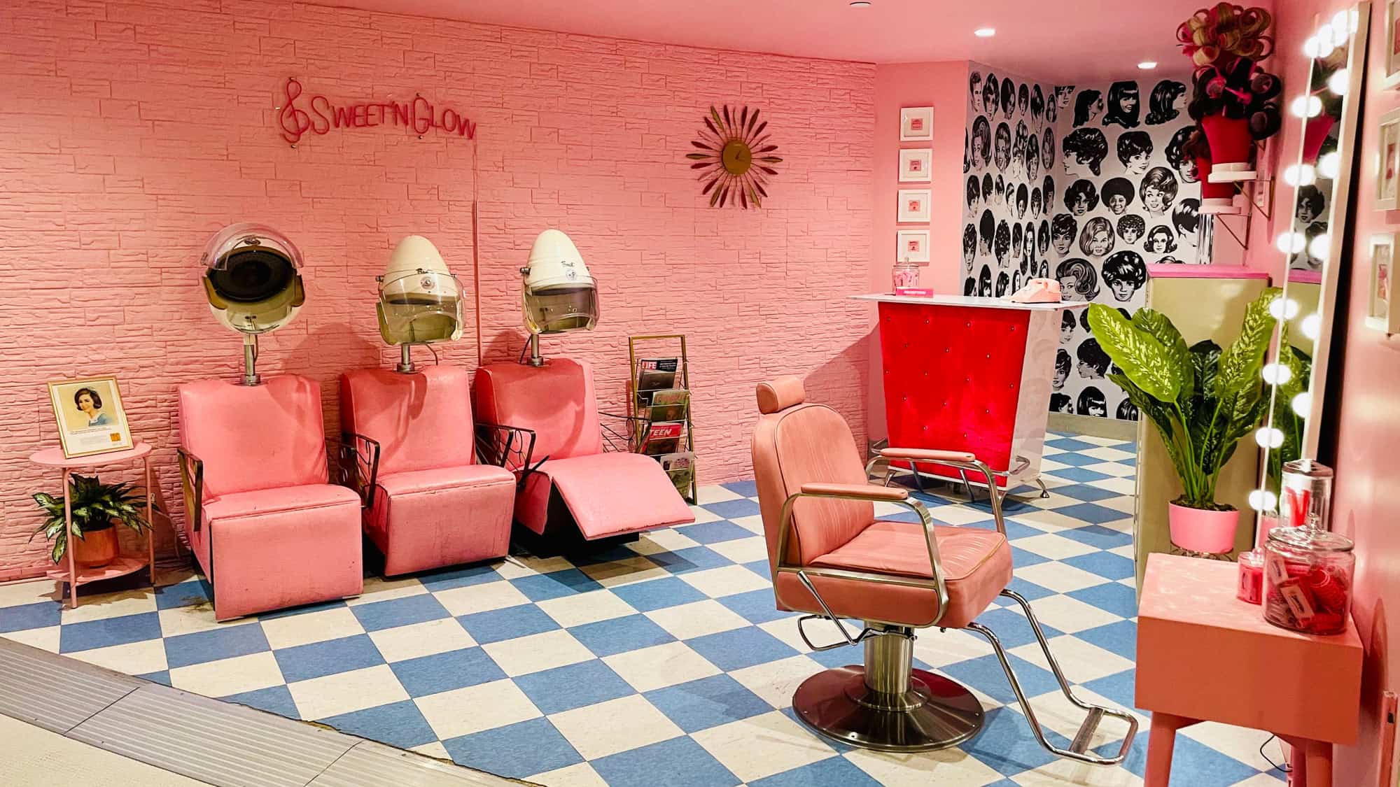 Retro-themed beauty salon with pink walls, blue and white checkerboard flooring, vintage hair dryers, and a neon sign reading 'SWEET'N GLOW.'